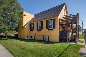 Um edifício amarelo com telhado inclinado, persianas pretas e escadaria de madeira que leva a uma porta.