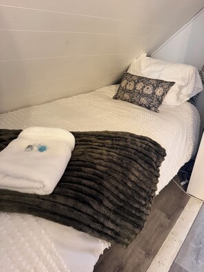 Uma cama de solteiro com edredom branco, manta marrom escura e almofadas estampadas.