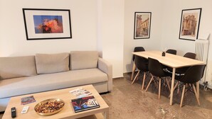 En moderne stue med en beige sofa, et træbord med magasiner og en skål med snacks, et spiseområde med et træbord og sorte stole, samt billeder i rammer på væggene.