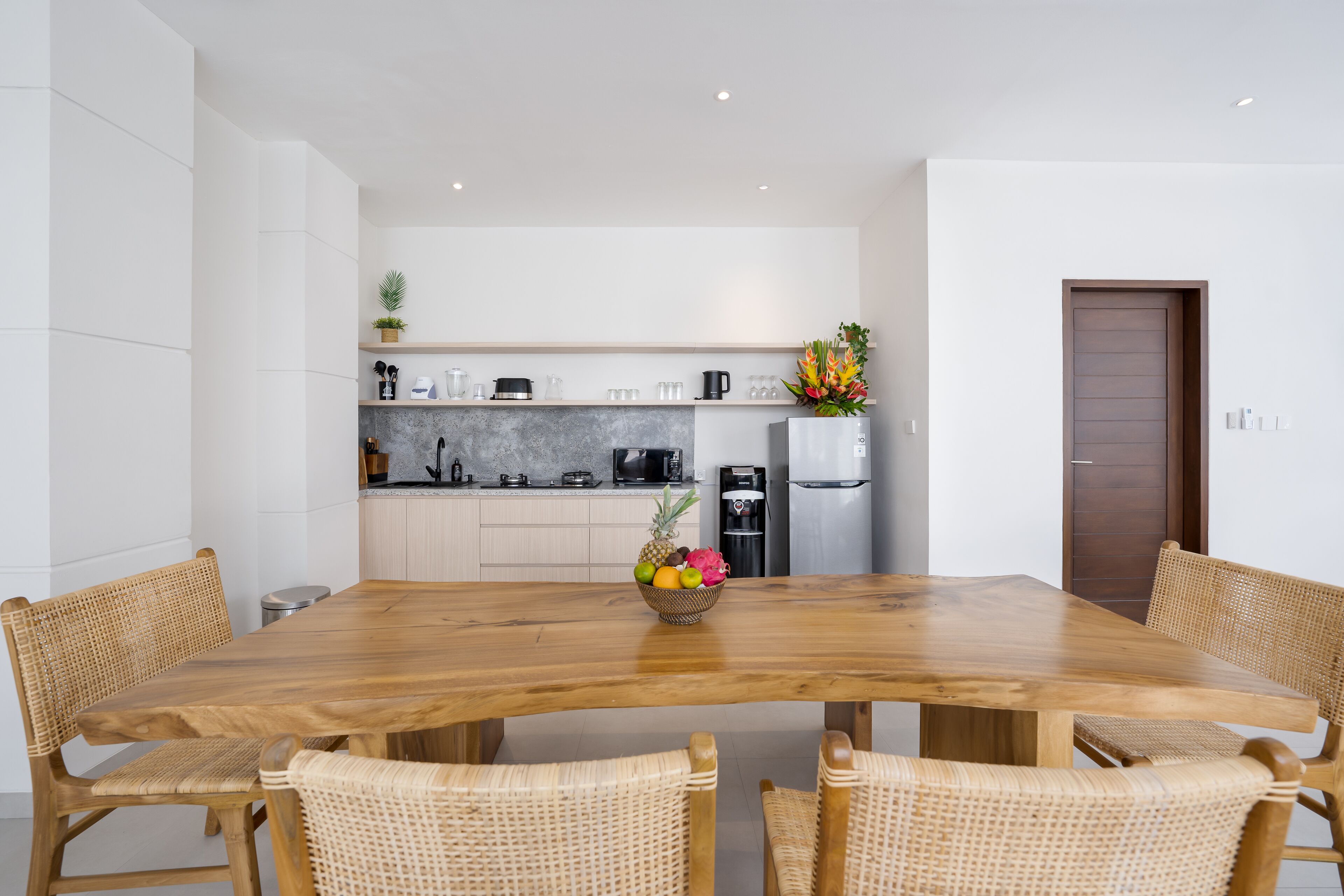 A modern kitchen with white cabinets, a wooden dining table, wicker chairs, and a fruit bowl.