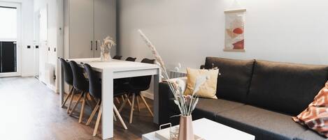 Uma sala de estar moderna com um sofá cinzento, uma mesa de centro branca e uma área de jantar com uma mesa branca e cadeiras pretas.