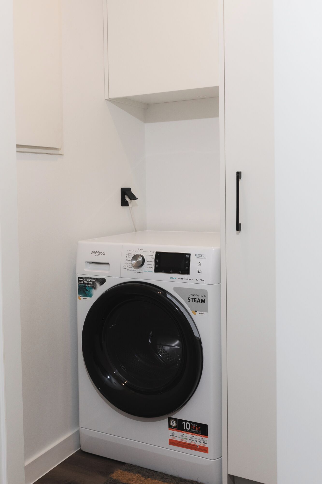 Un lave-linge et un sèche-linge Whirlpool, installés dans une buanderie blanche, avec une poignée de porte et un luminaire fixé au mur.