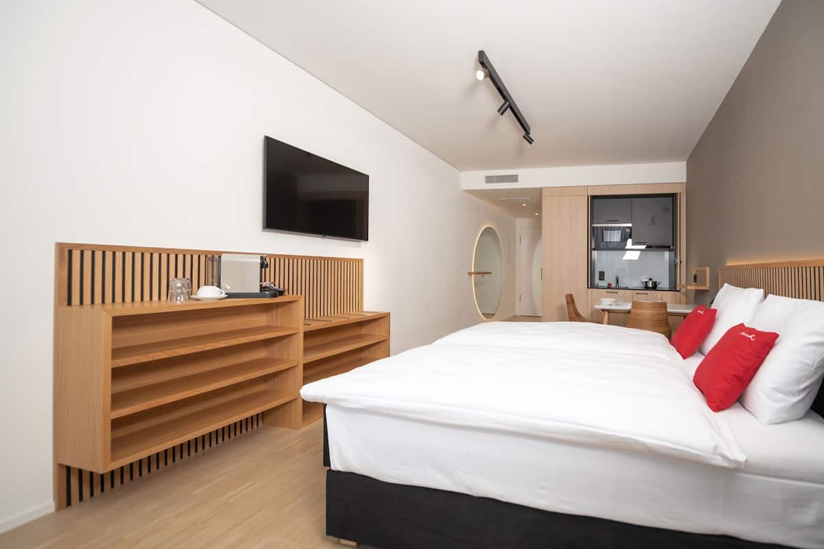 Ein modernes Hotelzimmer mit einem großen Bett, einem Holz-Kopfteil, eingebauten Regalen, einem Flachbildfernseher und einer kleinen Küche im Hintergrund.