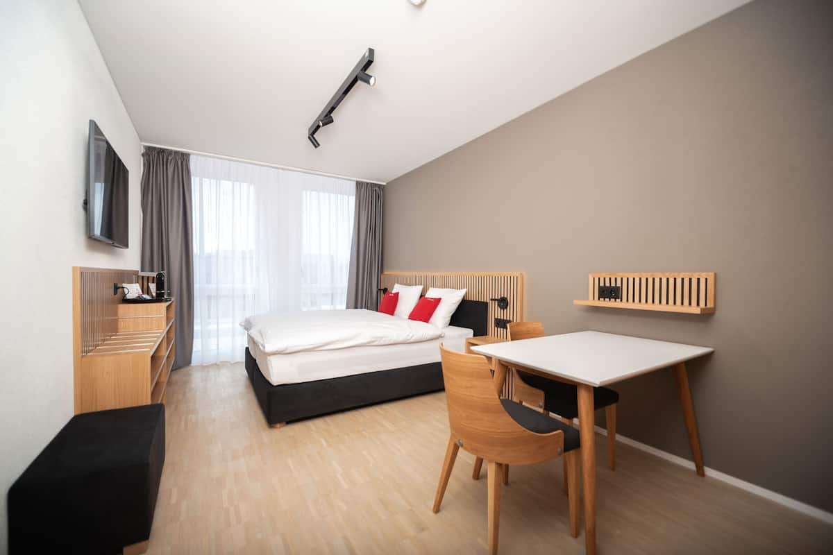 Ein modernes Hotelzimmer mit Bett, Nachttisch, Sessel, Schreibtisch und einem an der Wand befestigten Fernseher.