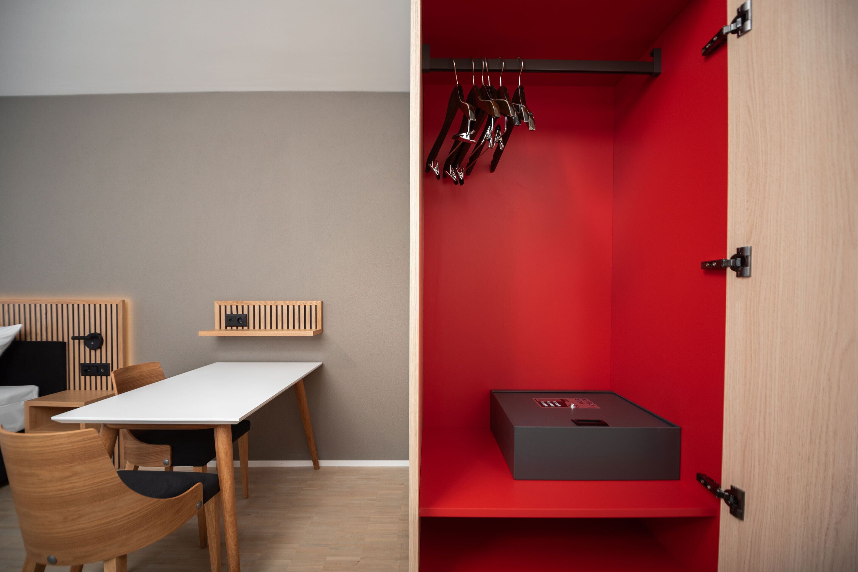 A room with a wooden table and chairs, a red accent wall, and a black box on a shelf.