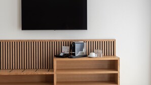 A modern hotel room with a flat-screen TV mounted on the wall, a wooden bench with a coffee machine, and a glass set on the bench.