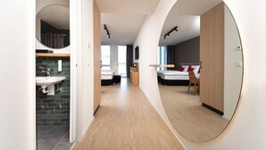 A modern hotel room with a wooden floor, a large mirror, a bed with white bedding, a bedside table, and a small seating area.
