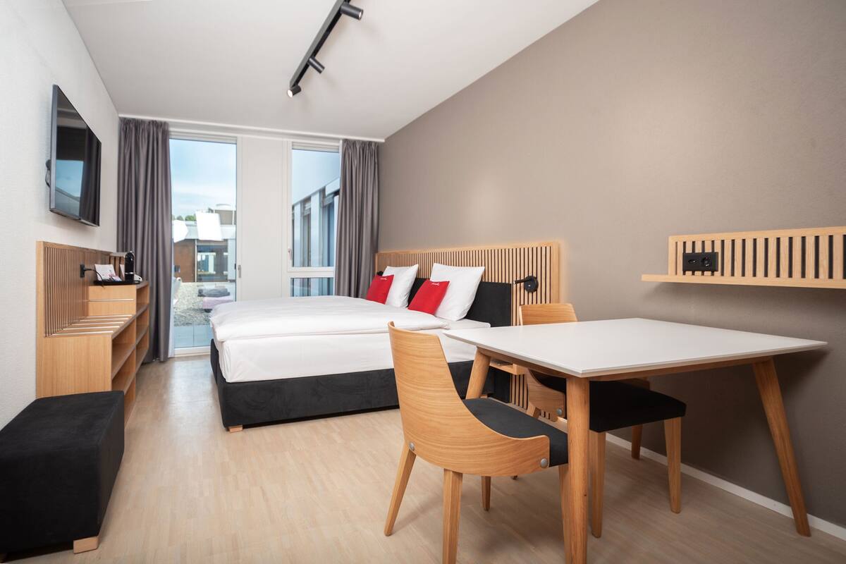 Ein modernes Hotelzimmer mit einem Bett, einem Schreibtisch, einem Sessel und einem an der Wand montierten Fernseher.
