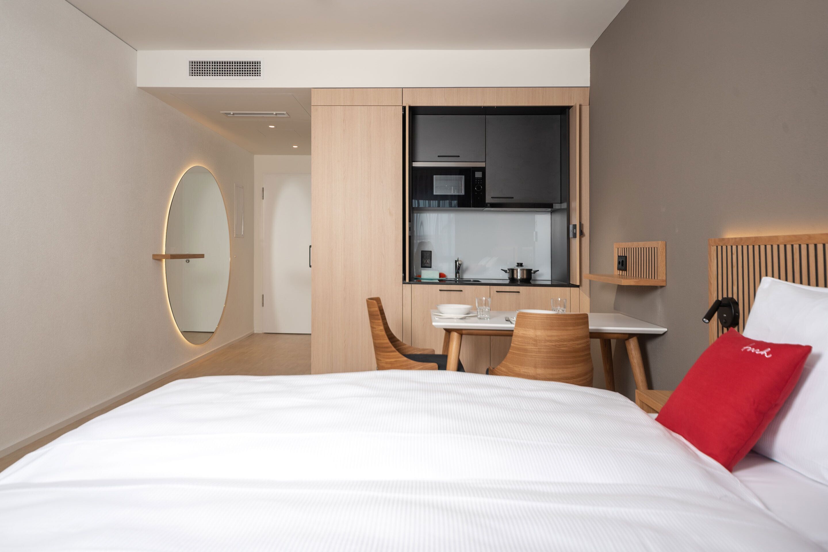 Ein modernes Hotelzimmer mit einem großen Bett, einem Essbereich mit Tisch und Stühlen, einer Küchenzeile mit Mikrowelle und Herd sowie einem runden Spiegel an der Wand.