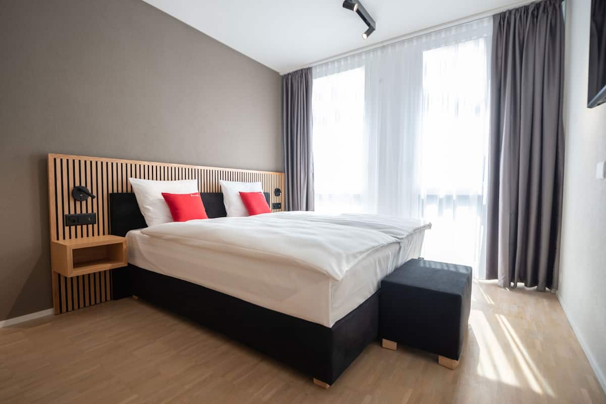 Ein modernes Schlafzimmer mit einem Holz-Kopfteil, einem großen Bett mit weißer Bettwäsche und roten Kissen, einem Nachttisch und einem schwarzen Hocker.