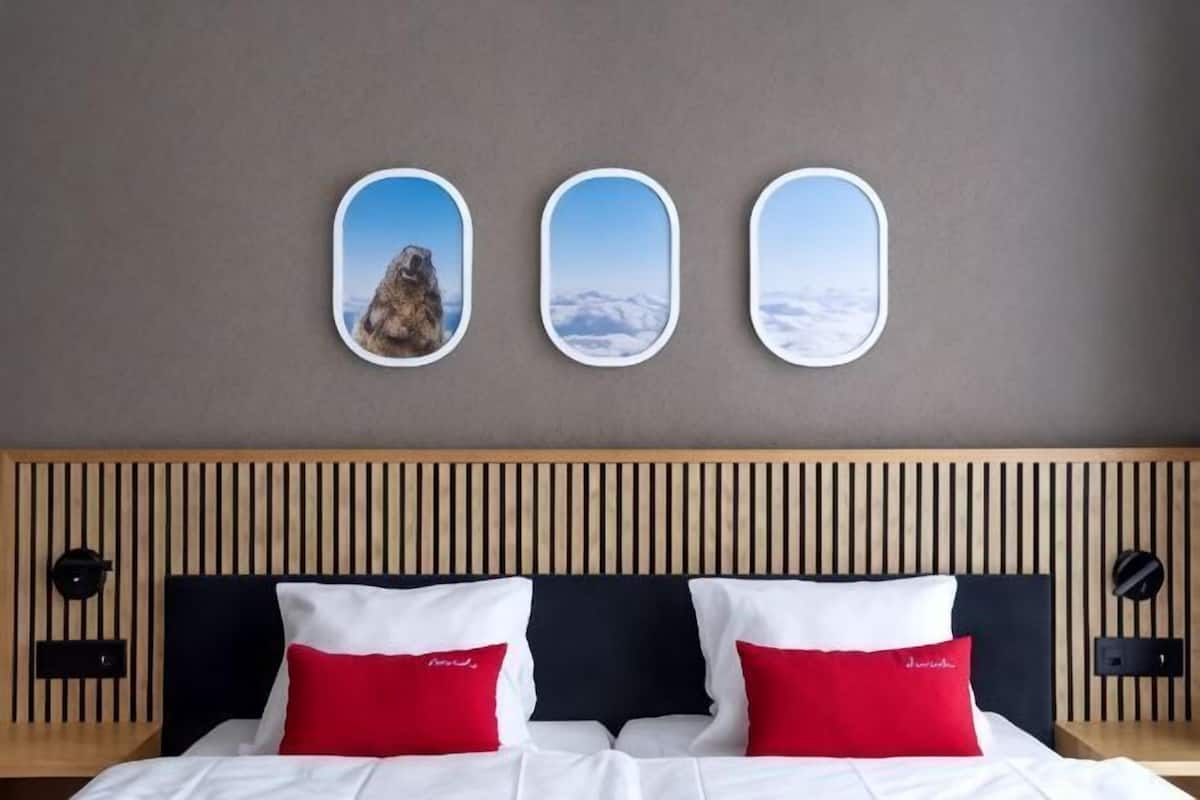 A hotel room with a bed, two red pillows, a wooden headboard, and three framed pictures on the wall.