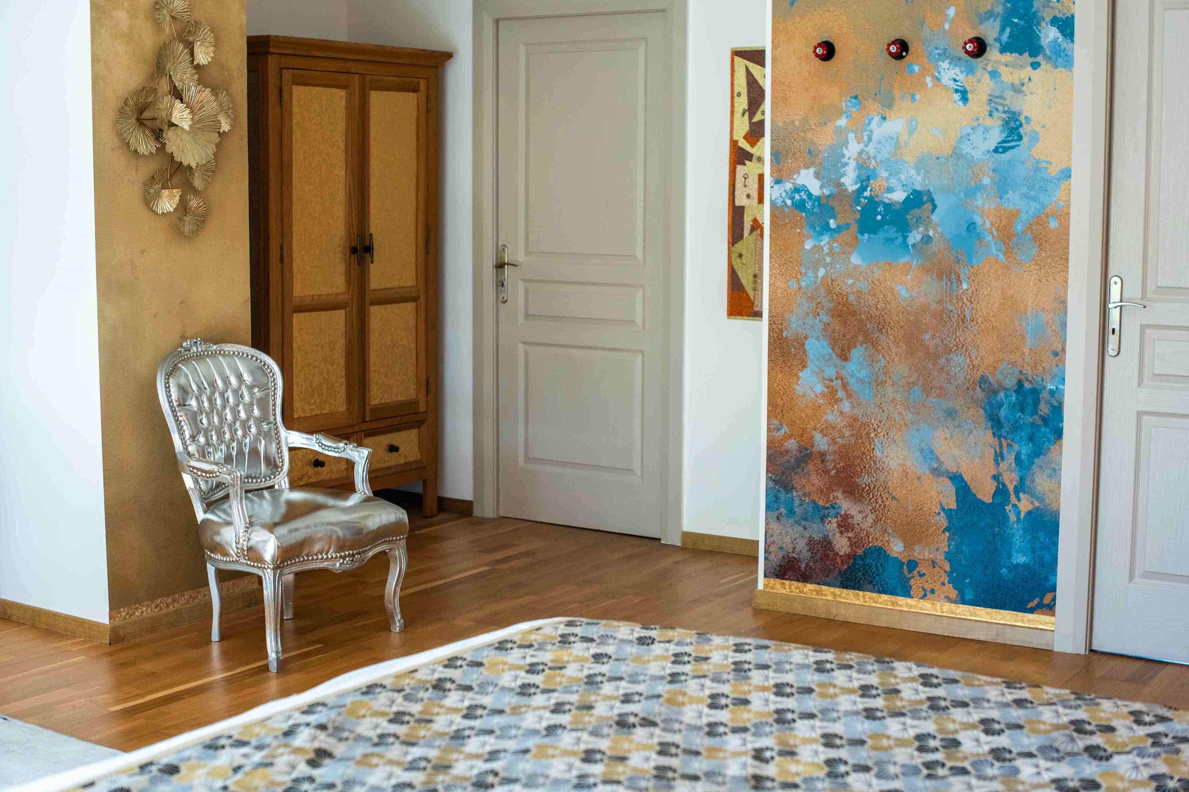 A room with a wooden floor, a white door, a silver chair, a patterned rug, and a wall with a blue and orange abstract painting.