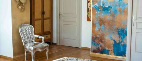 A room with a wooden floor, a white door, a silver chair, a patterned rug, and a wall with a blue and orange abstract painting.