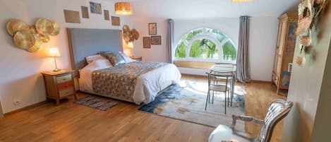Une chambre à coucher avec un grand lit, des tables de chevet, une chaise, une fenêtre donnant sur un beau paysage et des œuvres d’art décoratives au mur.