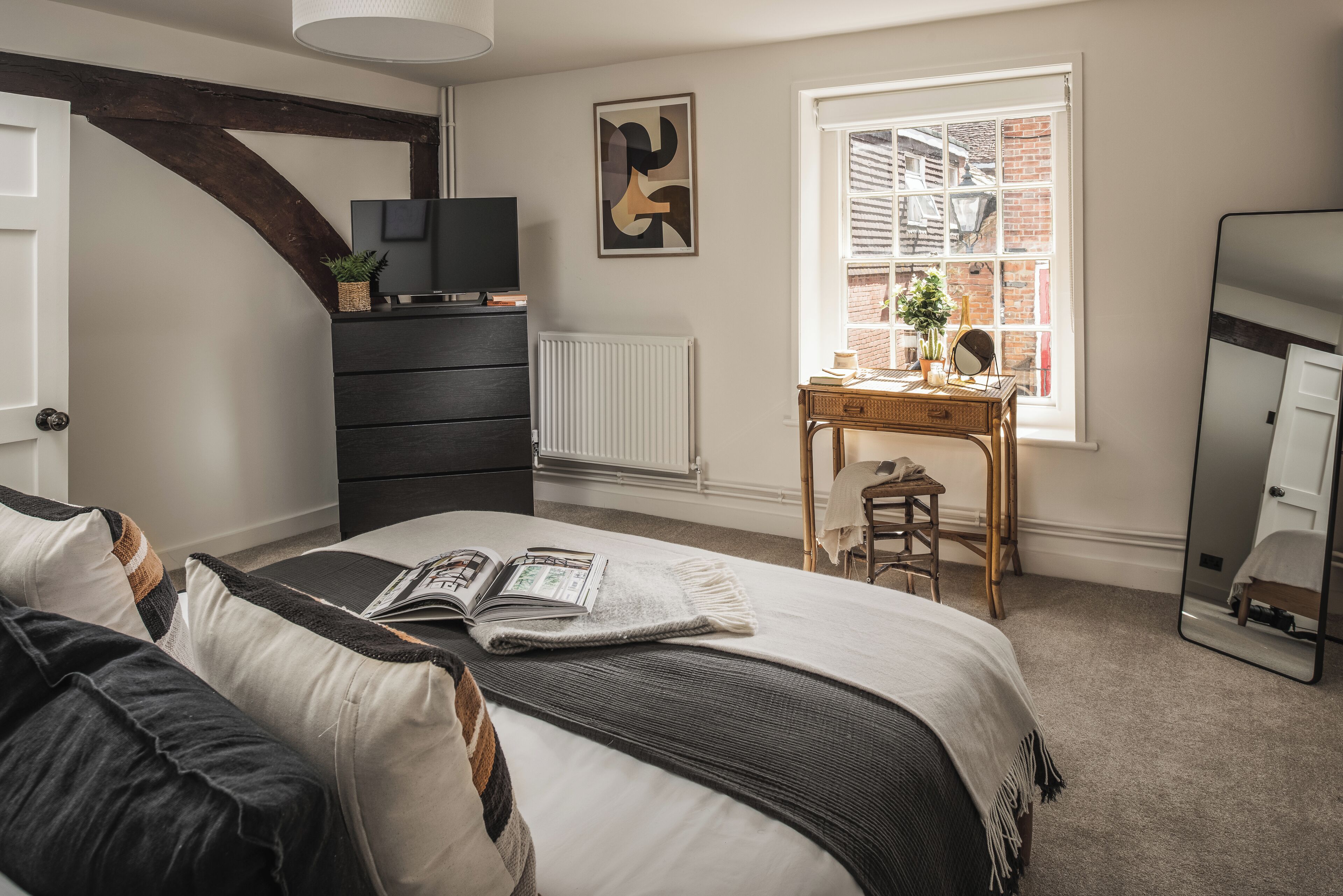 A bedroom with a bed, a dresser, a desk, a radiator, a window with a view of a brick building, and a mirror.