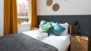 Una cama bien tendida con una colcha gris, un juego de toallas y artículos de aseo sobre la mesita de noche. Hay dos elementos decorativos en la pared sobre la cama y una ventana con cortinas a la izquierda.