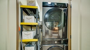 Uma lavanderia compacta com máquina de lavar e secar de abertura frontal, um carrinho utilitário amarelo com caixas organizadoras e um pequeno espelho na porta da secadora.