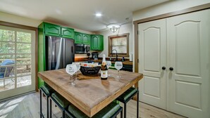 Uma cozinha com armários verdes, uma geladeira de aço inoxidável, uma mesa de madeira com uma garrafa e copos, e uma porta com maçaneta.