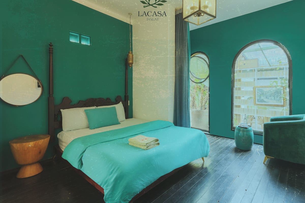 A teal-themed bedroom with a bed, a round mirror, a hanging lamp, and a window with a view.