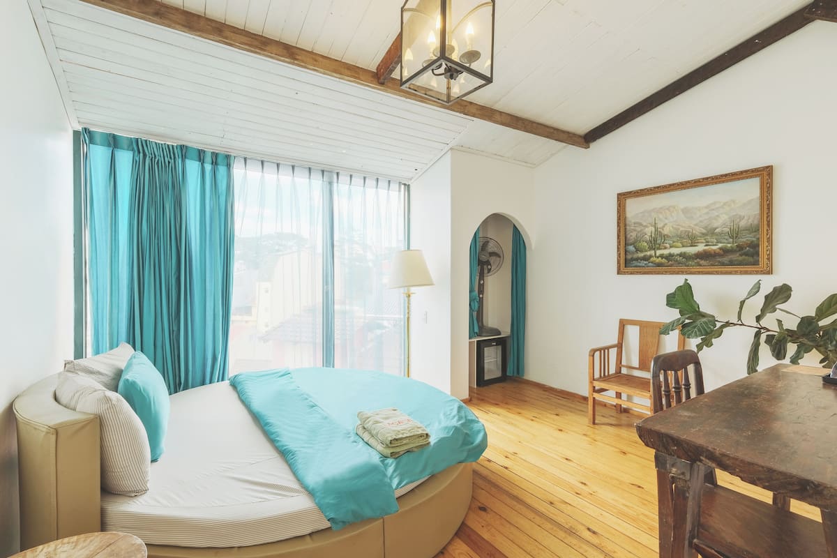 A room with a bed, wooden floor, a table, chairs, a lamp, and a painting on the wall.