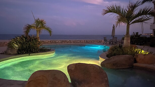 Una piscina con borde curvo, rodeada de rocas y palmeras, con vista al océano al atardecer.