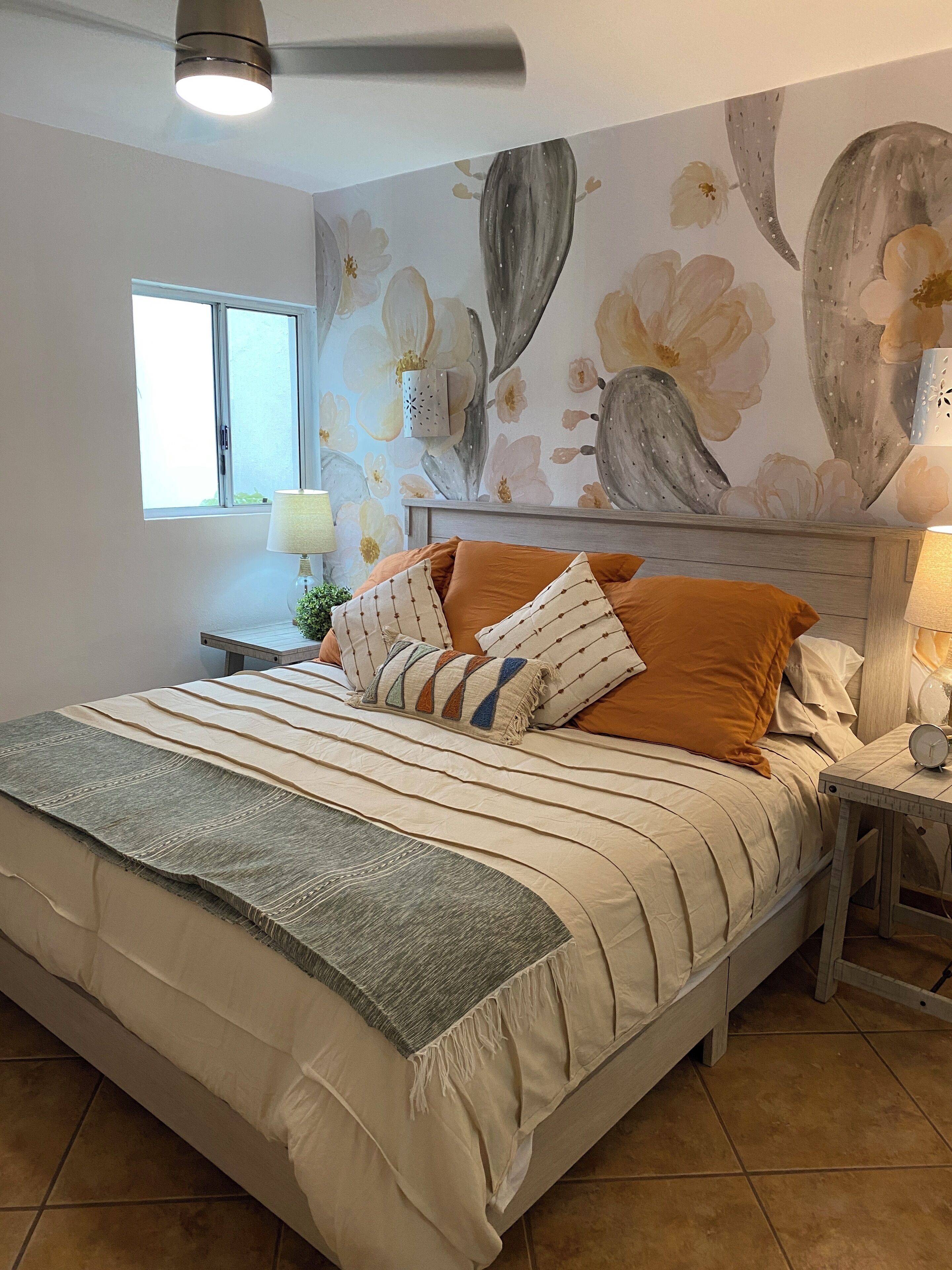 Een slaapkamer met een bed, nachtkastje, lamp, raam en een bloemen behang.