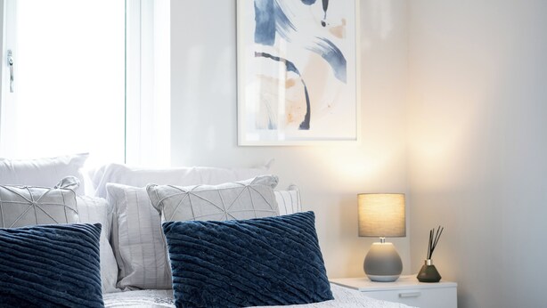 Un dormitorio con cama, almohadas, una mesita de noche, una lámpara y un cuadro abstracto colgado en la pared.