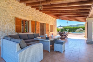 Un área de estar al aire libre con techo, mobiliario de mimbre, paredes de piedra y vistas a una piscina y vegetación.