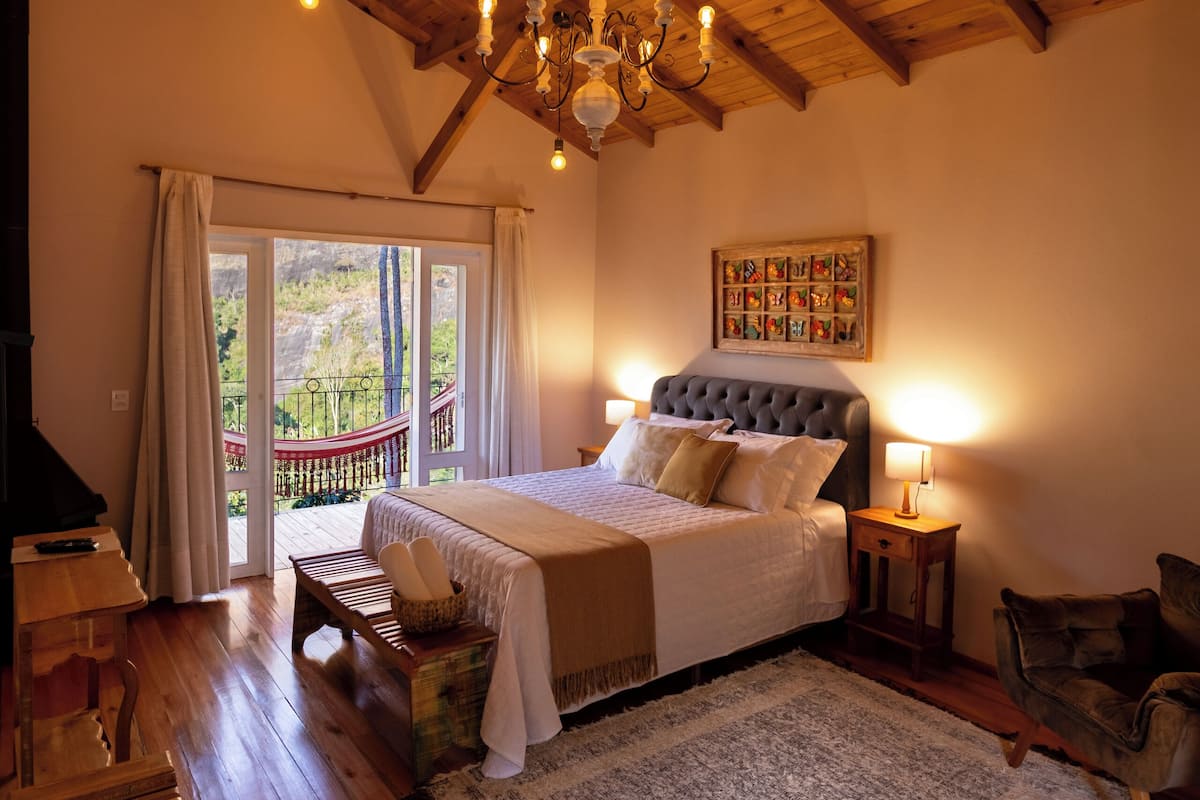 Een ruime slaapkamer met een groot bed, houten balken in het plafond, een kroonluchter en uitzicht door een glazen deur.