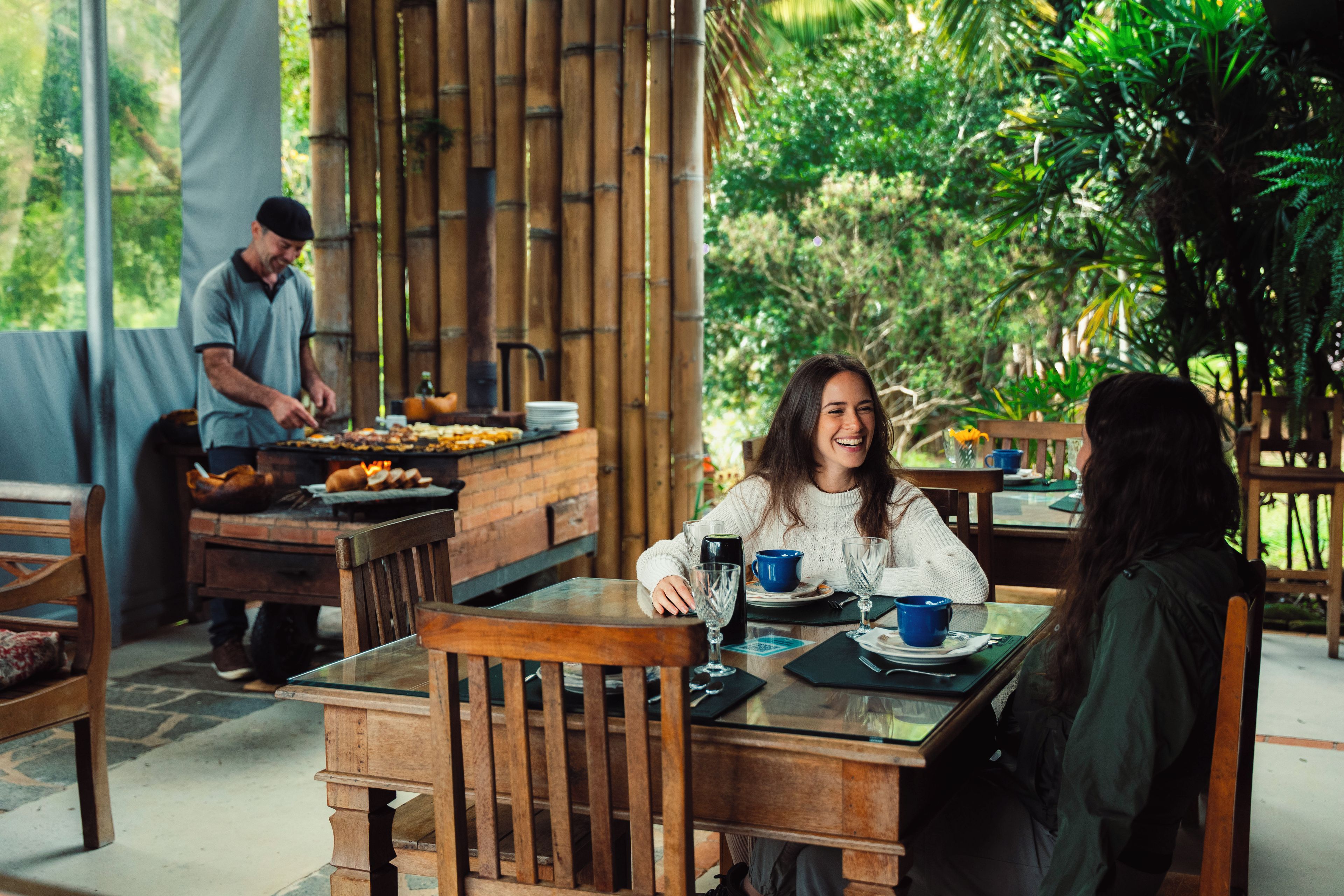 An outdoor dining area with wooden tables and chairs, a chef preparing food, and lush greenery in the background.