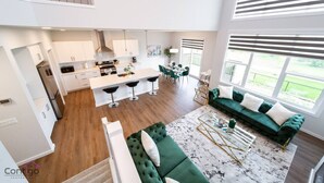 A modern kitchen with a central island, a dining area with chairs, a green sofa, and a view of the outdoors. - Orchards Retreat | AirCon | Sleeps 10 | Near Airpt (Edmonton)