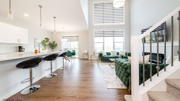 A modern kitchen with white cabinets, a bar area with stools, a green sofa, and a staircase. - Orchards Retreat | AirCon | Sleeps 10 | Near Airpt (Edmonton)