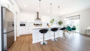 A modern kitchen with white cabinets, a stainless steel refrigerator, a central island with bar stools, and a large window providing natural light. - Orchards Retreat | AirCon | Sleeps 10 | Near Airpt (Edmonton)