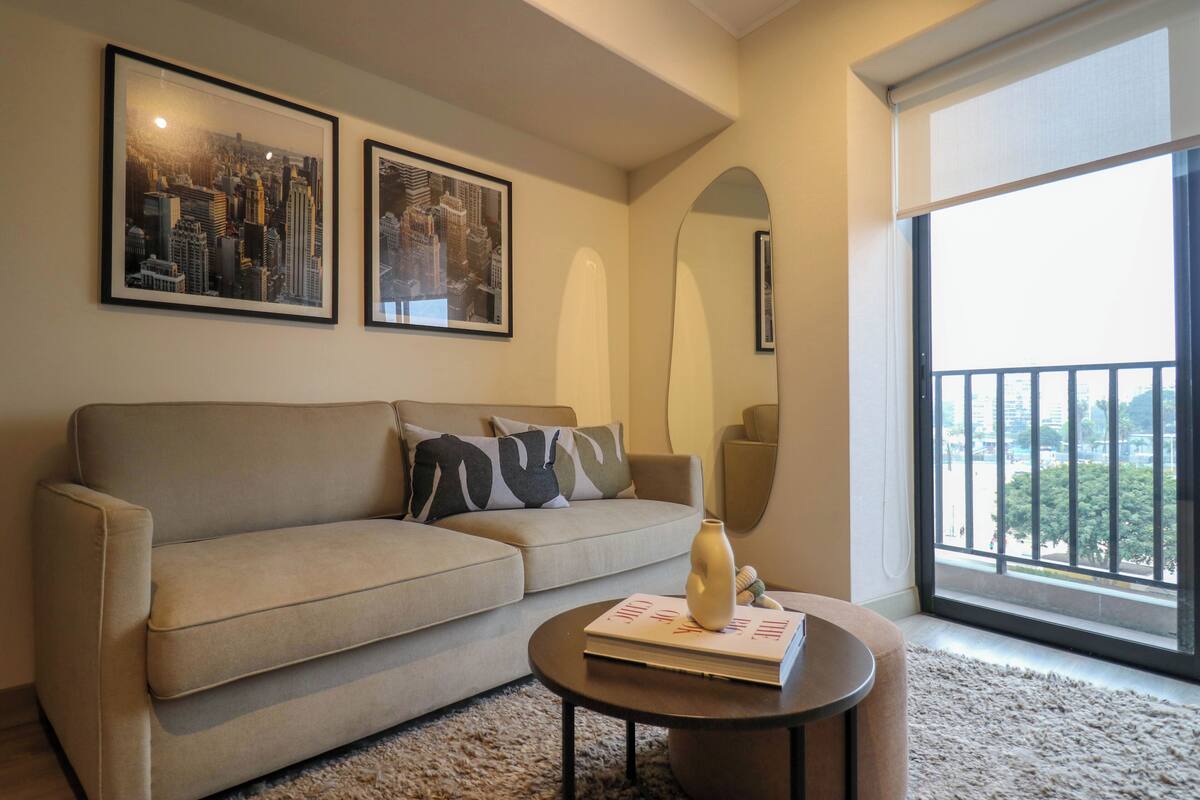 A living room with a beige sofa, two framed cityscape photographs, a round coffee table with a book and vase, and a large window with a view of buildings.