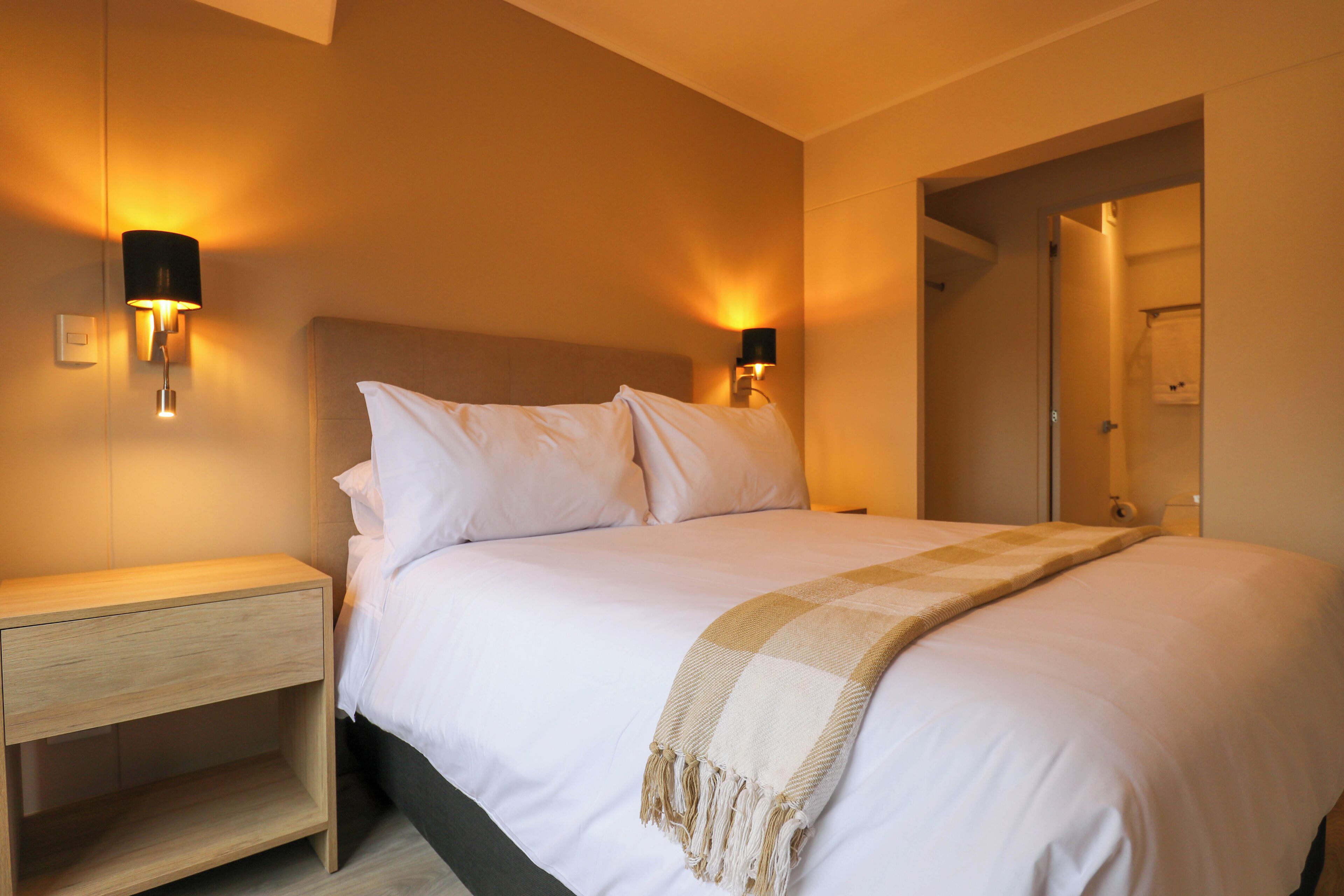 A modern hotel room with a large bed, two bedside lamps, a wooden nightstand, and a door leading to a bathroom.