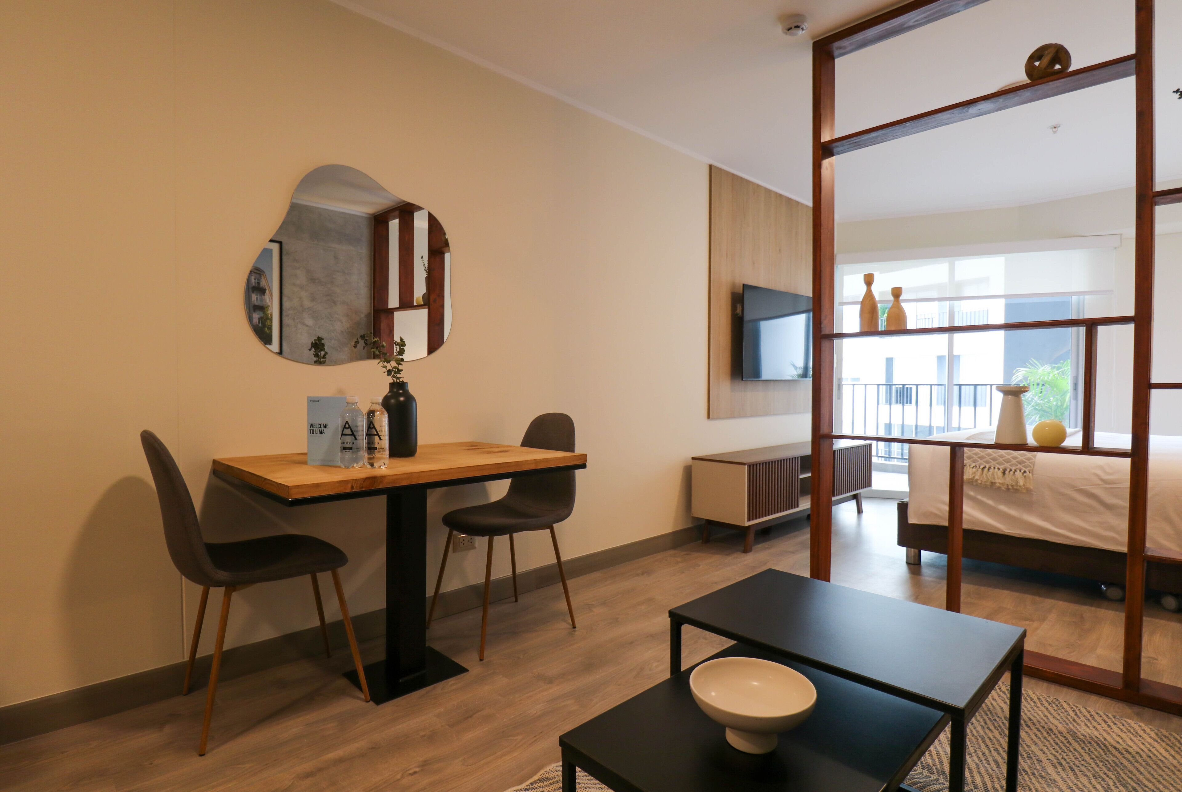 A modern hotel room with a wooden desk, two chairs, a small table with a bowl, a TV, a bed with a view, and a large mirror.