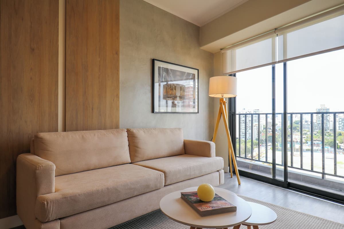 A modern living room with a beige sofa, a round coffee table with a book and a yellow object, a floor lamp, and a large window with a city view.