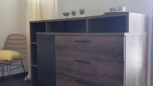 A room with a wooden cabinet with drawers and shelves, a yellow chair, and a window with curtains. - 1 Bedroom Apartment in Saint-francois (Saint-francois)