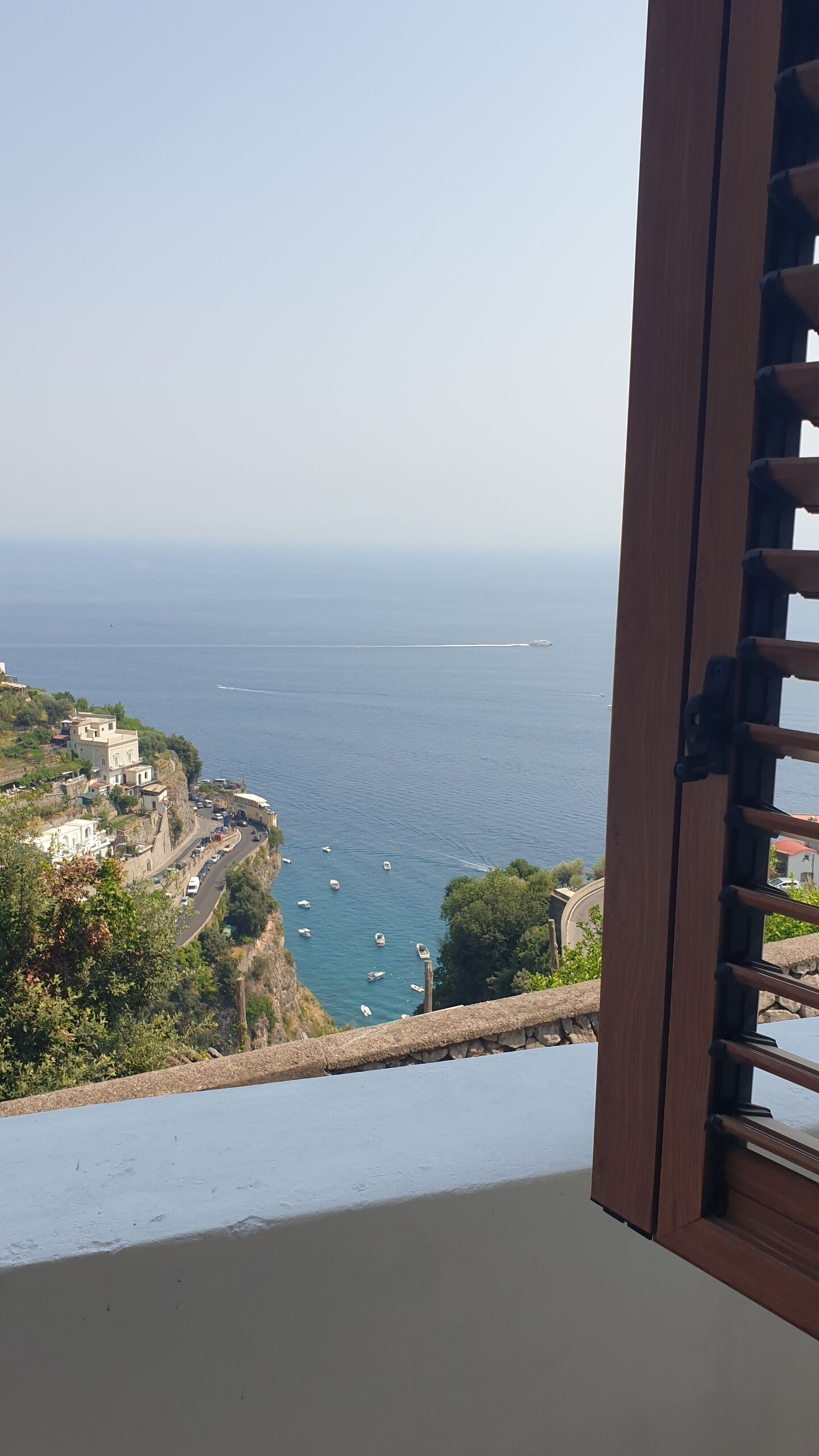 Une vue sur la côte, aperçue à travers une fenêtre ornée d’un volet en bois, avec une mer où naviguent des bateaux, une route et des bâtiments sur une colline.