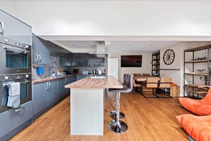 A modern kitchen with a central island, built-in appliances, and a dining area with a wooden table and chairs. - Stunning 5 Bed House in Leeds Contractors Welcome (Leeds)