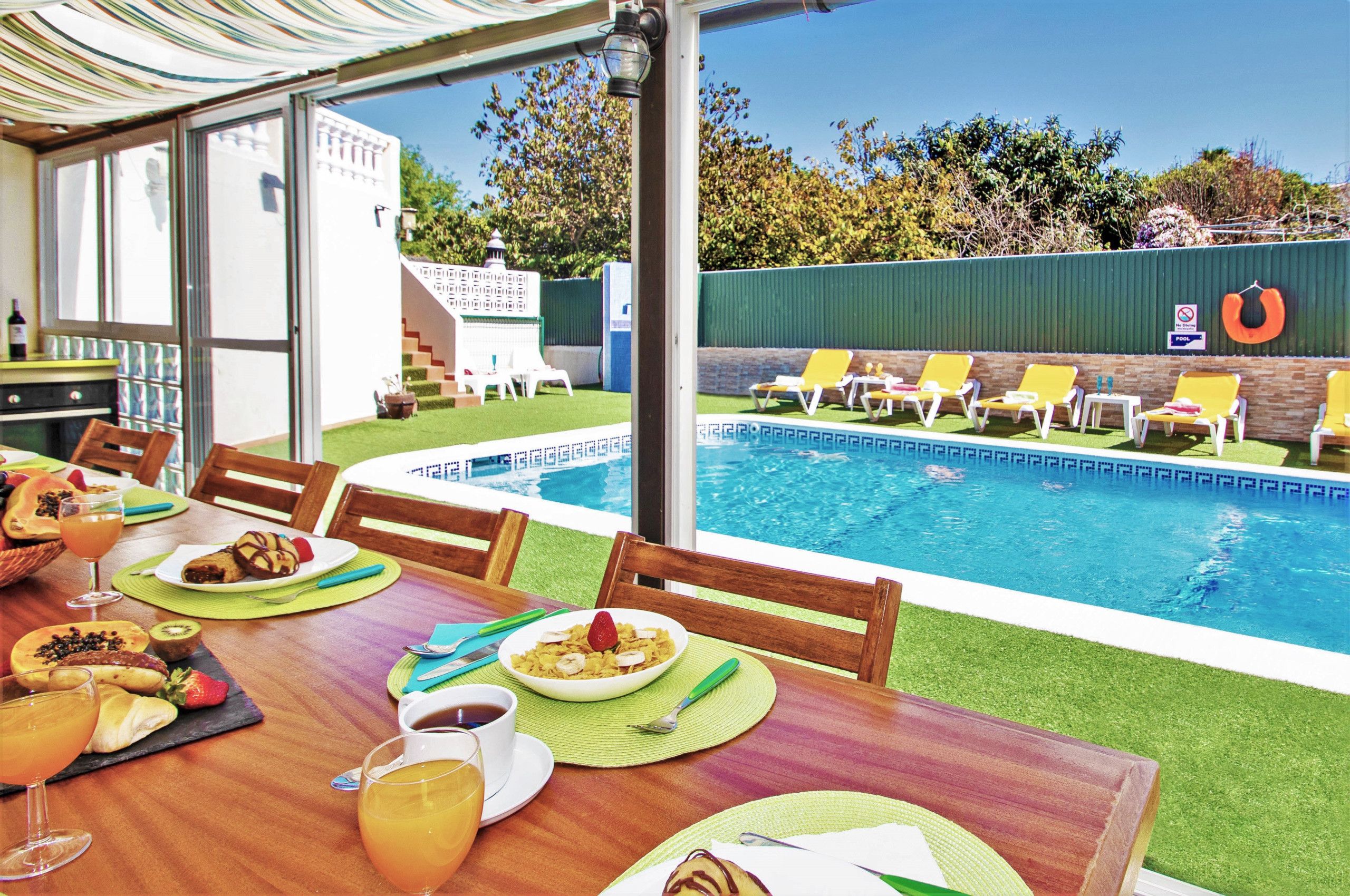 An outdoor dining area with a pool view, a table set with food and drinks, and a poolside lounge area with chairs and a no diving sign.