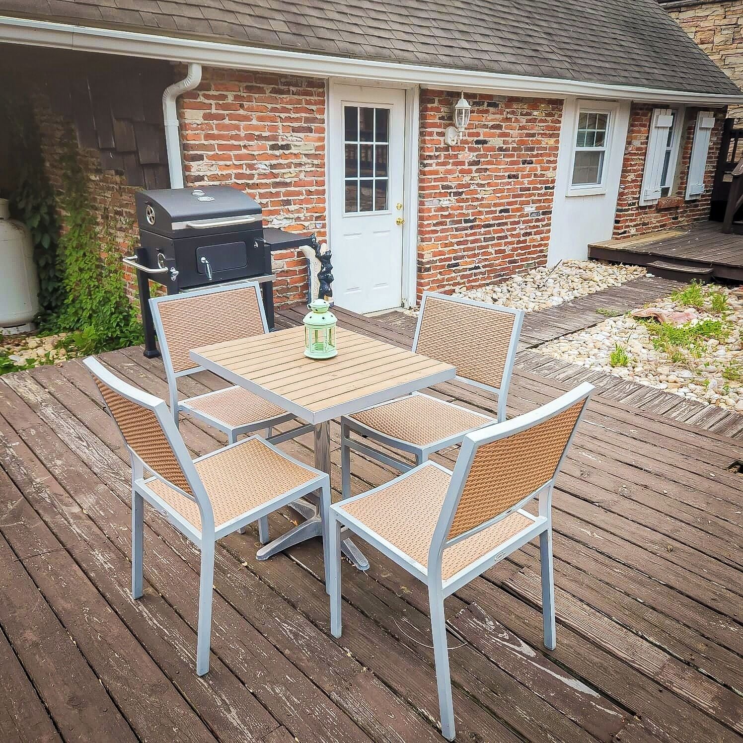 En terrasse med et bord og stole, en grill og et murstenshus i baggrunden.