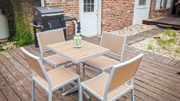 En terrasse med et bord og stole, en grill og et murstenshus i baggrunden.
