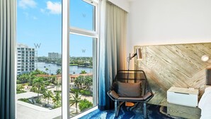 A hotel room with a large window offering a view of a river and cityscape, a modern chair, a bedside table, and a headboard with a geometric pattern.