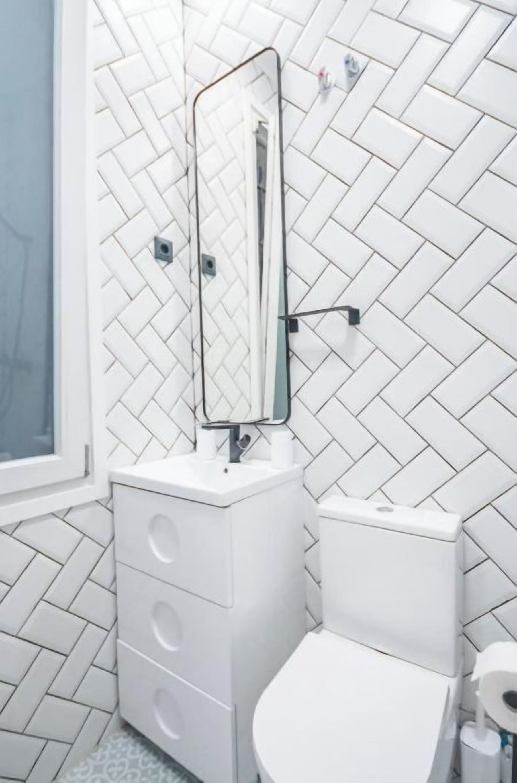 Un baño con baldosas blancas de patrón chevron, un tocador blanco con espejo y un inodoro.