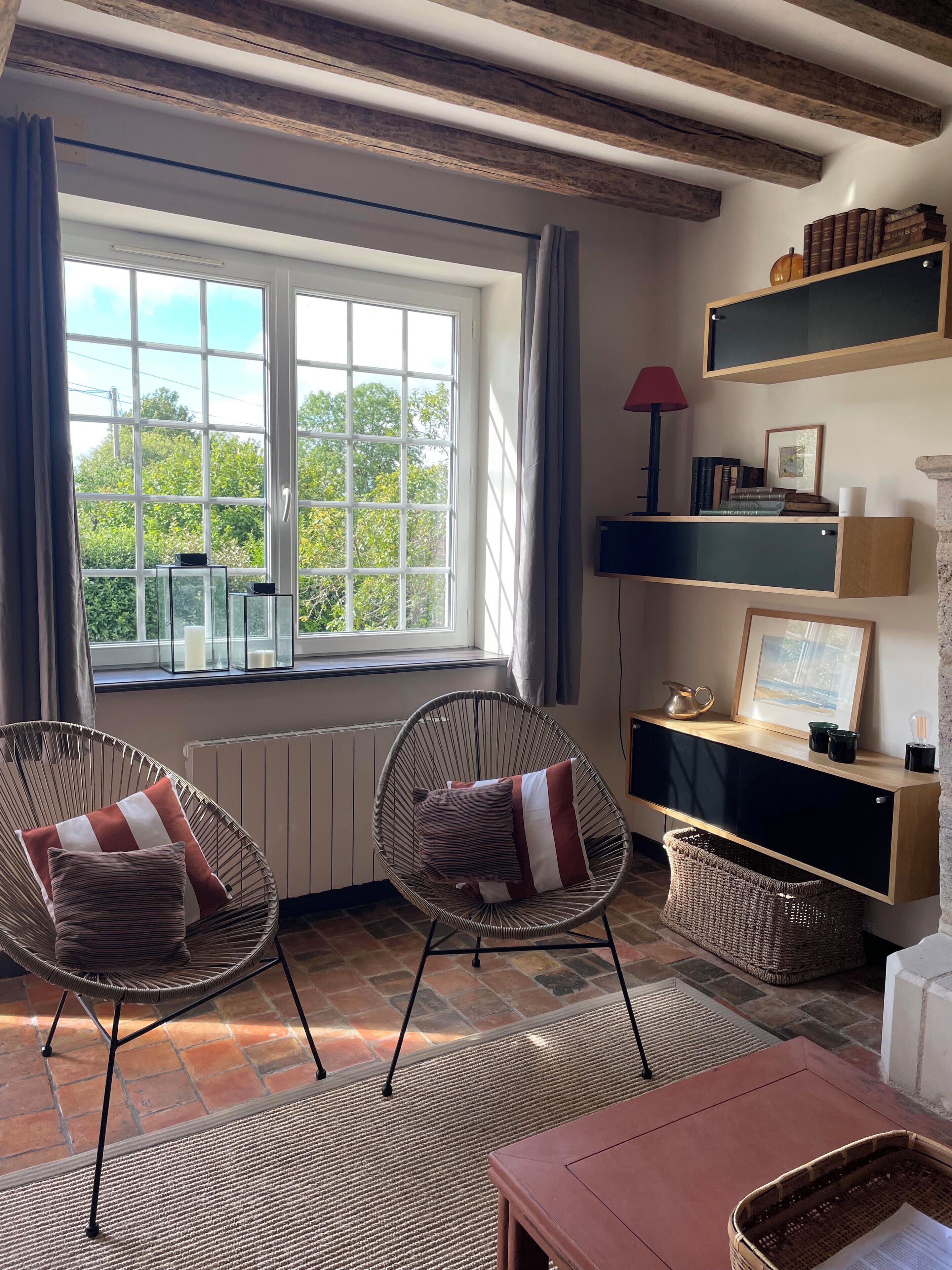 A room with two wicker chairs, a wooden table, a radiator, a window with curtains, a shelf with books and decorative items, and a view of greenery outside.