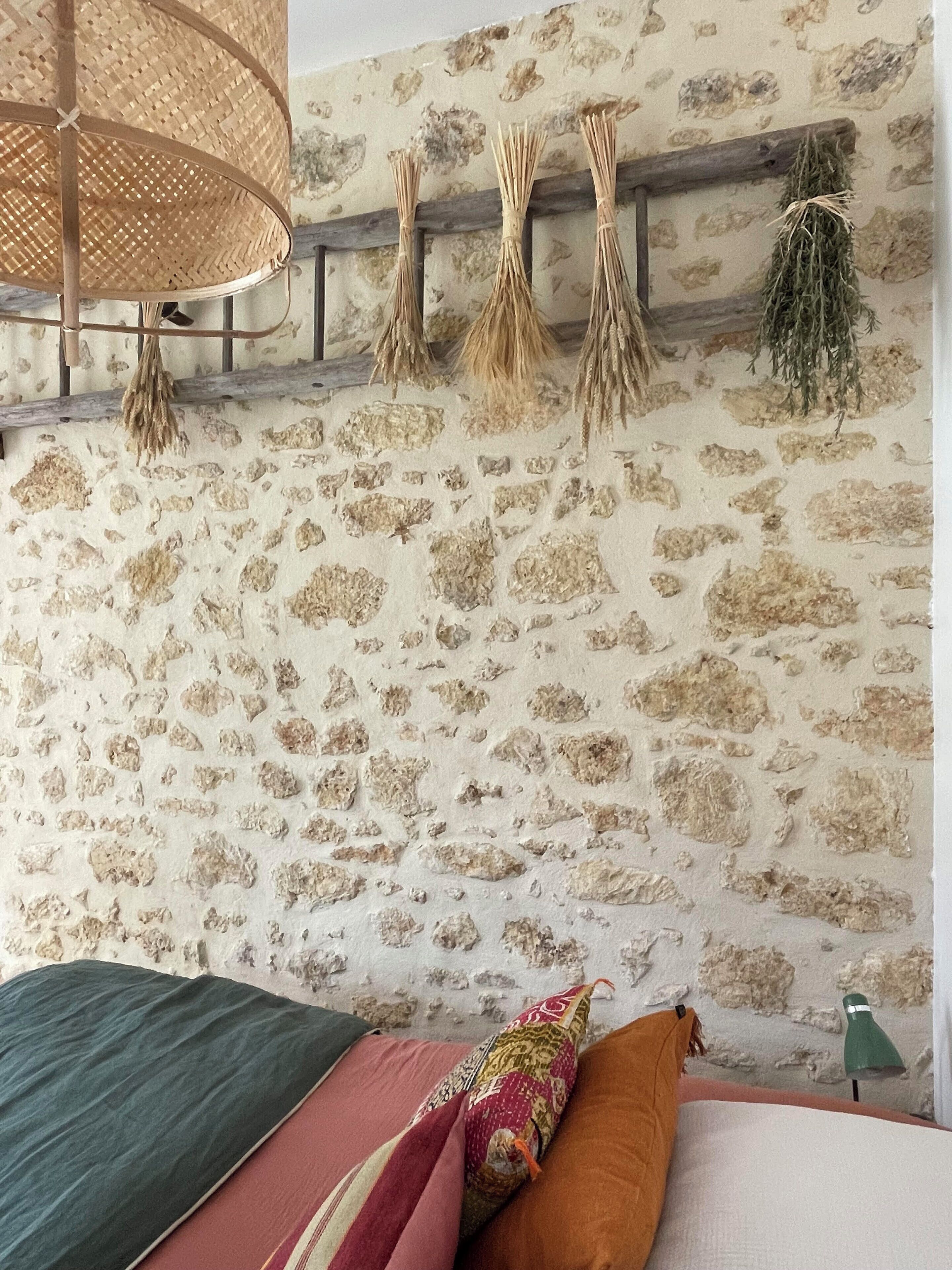 A bedroom with a stone wall, a bed with multiple pillows, a hanging light fixture, and a wooden shelf with dried plants.