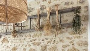 Un dormitorio con pared de piedra, una cama con muchos almohadones, una lámpara colgante y una repisa de madera con plantas secas.