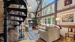 A spacious living area with a stone fireplace, wooden beams, and large windows offering natural light.