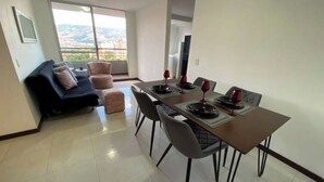 Uma sala de jantar com mesa posta para quatro pessoas, um sofá e vista para a paisagem urbana através de uma grande janela.